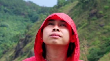 Dağın tepesinde duran ve manzaranın tadını çıkaran kırmızı su geçirmez bir ceket giyen mutlu bir Asyalı kadın yürüyüşçünün portresi..