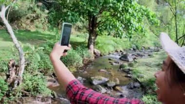 Güzel bir Asyalı kadın dağ ormanındaki bir derede tahta bir köprüde oturur ve akıllı telefonuyla video görüşmesi yapar..