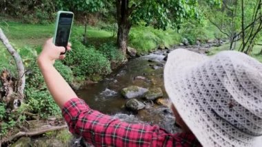 Güzel bir Asyalı kadın dağ ormanındaki bir derede tahta bir köprüde oturur ve akıllı telefonuyla selfie çeker. Tatil yaşam tarzı ve maceralar.