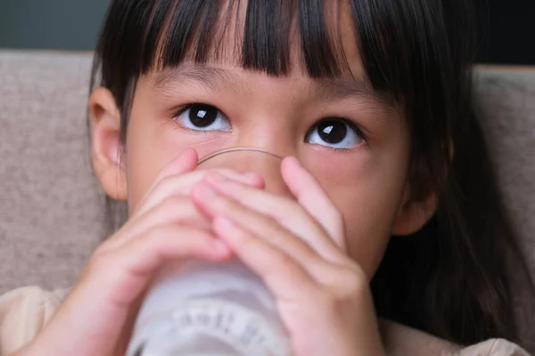 自宅のソファに座っているミルクのガラスを持ってかわいいアジアの少女の肖像画 笑顔で家にいる小さな女の子 ミルクを飲みながらカメラを見て幸せを感じる — ストック写真