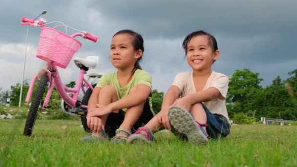 公園の自転車の近くの芝生に座っている幸せなかわいい女の子と彼女の妹 子供たちは自転車で休んでいる 子供のための健康的な夏の活動 — ストック動画