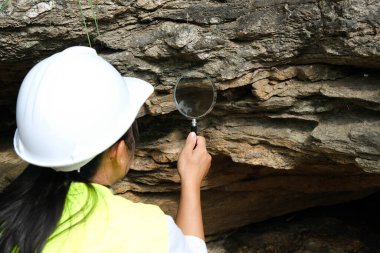 Asian female geologist researcher analyzing rocks with a magnifying glass in a natural park. Exploration Geologist in the Field. Stone and ecology concept. clipart