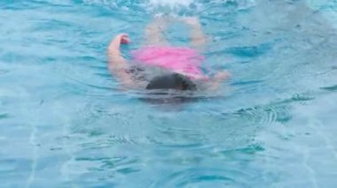 Happy little girl is swimming and playing in outdoor swimming pool in a tropical resort during family summer vacation. Girl learning to dive and swim. Healthy Summer Activities for Kids.