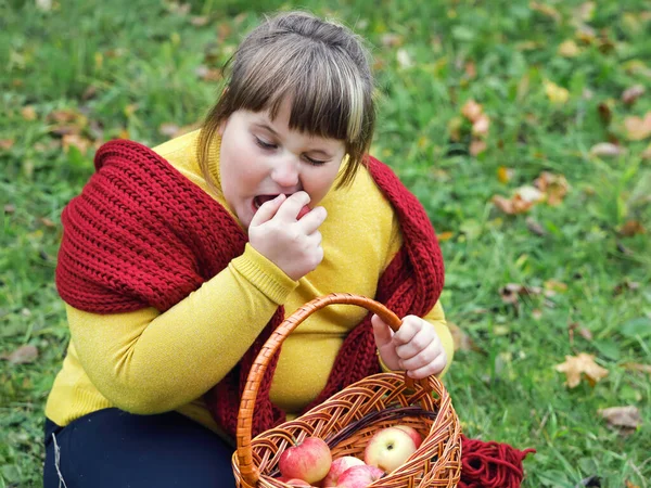 赤いスカーフを持つ肥満の女の子は草の上に座って 秋にリンゴとバスケットを保持し リンゴをかむ — ストック写真