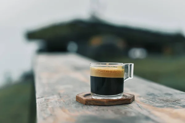 A clear cup of coffee is placed on a brown lacquered wooden surface with a naturally blurred background.