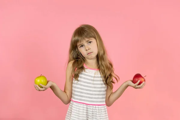 一个脸色苍白的小女孩正试图在一个绿色苹果和一个红色苹果之间做出选择 小女孩不知道选哪个苹果 一个手里拿着苹果的小女孩站在一个彩色的背景上 — 图库照片