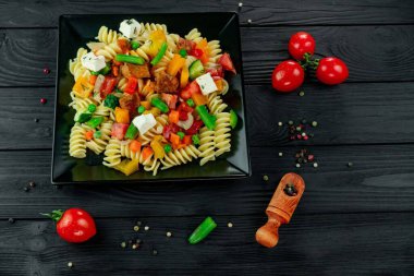 Sebzeli makarna, bezelye, tatlı biber, domates, sos, siyah ahşap arka planda mozzarella peyniri. Karanlık bir arka planda sebzeli ve baharatlı İtalyan makarnası..