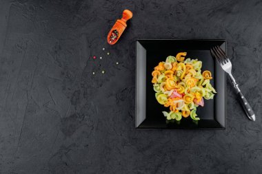 Siyah beton arka planda çok renkli makarna. Düz yatıyordu. Üst Manzara. Çok renkli farfalle, seçici odaklı, stüdyoda çekilmiş siyah bir kase..