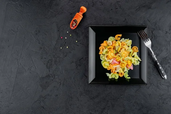 Siyah beton arka planda çok renkli makarna. Düz yatıyordu. Üst Manzara. Çok renkli farfalle, seçici odaklı, stüdyoda çekilmiş siyah bir kase..