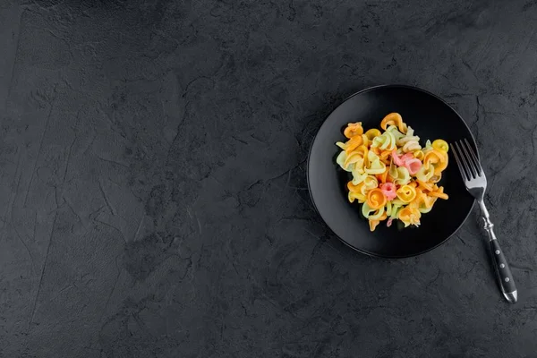 Siyah beton arka planda çok renkli makarna. Düz yatıyordu. Üst Manzara. Çok renkli farfalle, seçici odaklı, stüdyoda çekilmiş siyah bir kase..