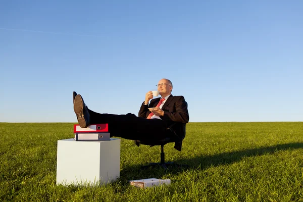 Бізнесмен, насолоджуючись кава на стілець в трав'янистих області проти неба — стокове фото
