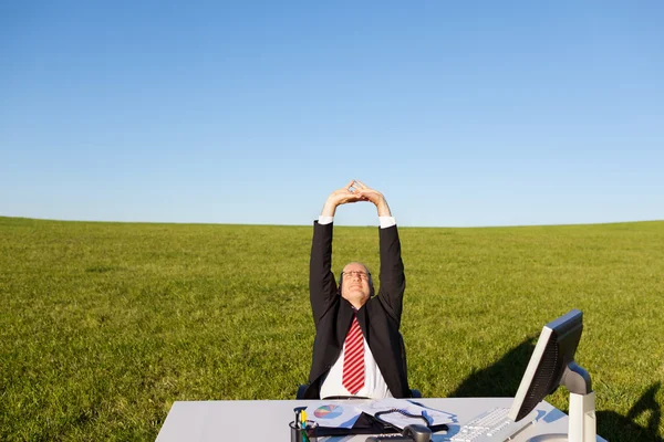 Affärsman stretching vid skrivbord på gräsbevuxna fält mot himlen — Stockfoto