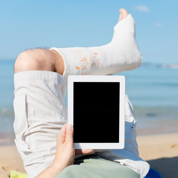 Man med gips ligga på stranden med ipad — Stockfoto