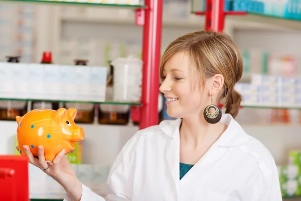 Smiling pharmacist looking at piggy bank — Stock Photo, Image