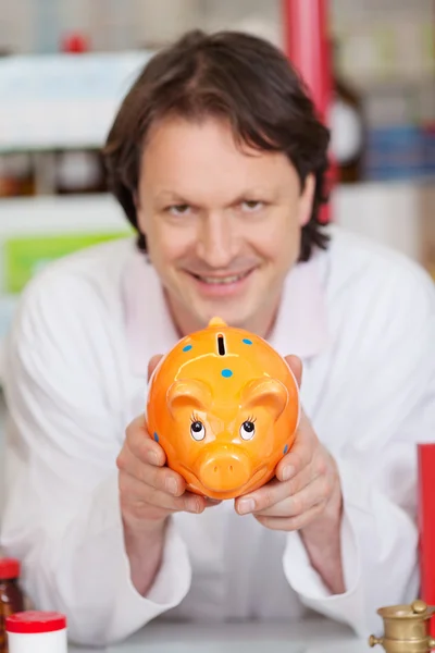 Confident pharmacist showing piggy bank — Stock Photo, Image