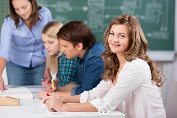 Adolescente chica sonriendo con profesor asistiendo compañeros de clase en el escritorio —  Fotos de Stock