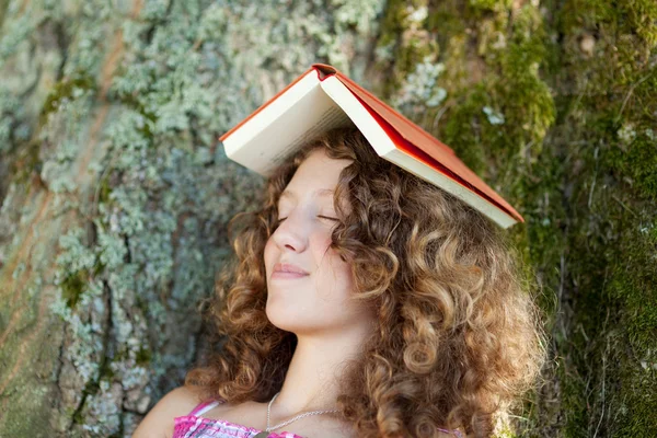 Meisje met boek over hoofd dutten op boomstam — Stockfoto