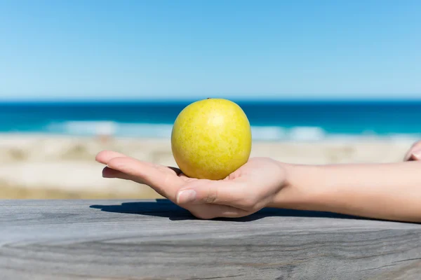 Kvinnans hand innehav grönt äpple på trä på stranden — Stockfoto