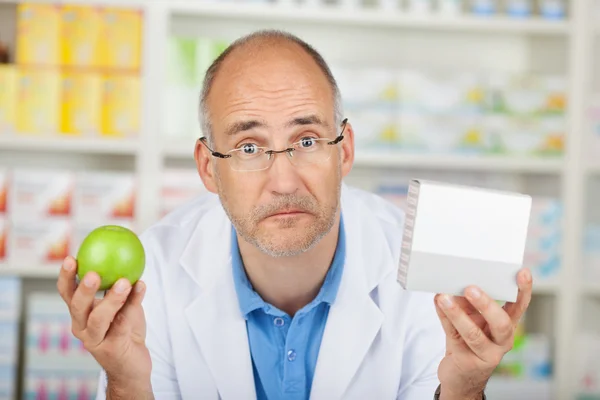 Farmacéutico preguntándose mientras sostiene Apple y la caja de la medicina —  Fotos de Stock