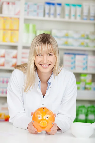 Apotheker hält Sparschwein, während er sich an Apothekentheke lehnt — Stockfoto