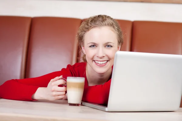 Kobieta z laptopa trzyma kubek latte w kawiarni — Zdjęcie stockowe