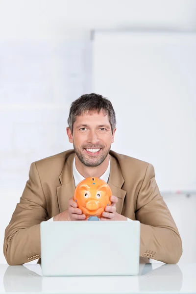 Бизнесмен со свиным банком и ноутбуком в офисе — стоковое фото