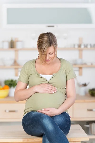 Femme enceinte avec les mains sur le ventre assis sur le comptoir de cuisine — Photo