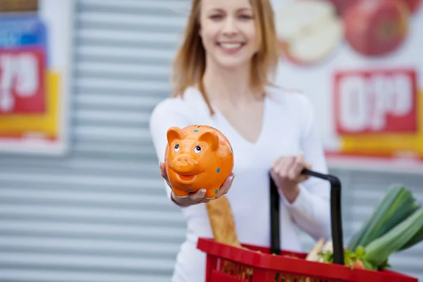 Mujer mostrando piggybank mientras sostiene cesta de la compra — Foto de Stock