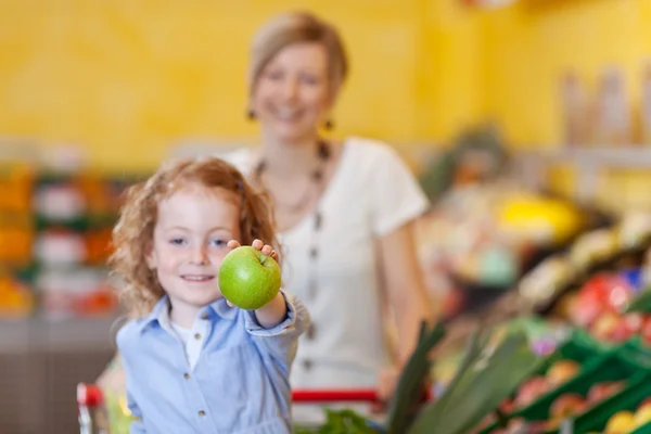 Chica mostrando manzana con madre en segundo plano en la tienda —  Fotos de Stock
