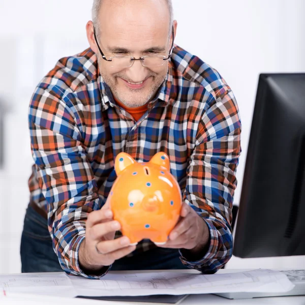 Älterer Mann lächelt im Büro auf ein Sparschwein — Stockfoto