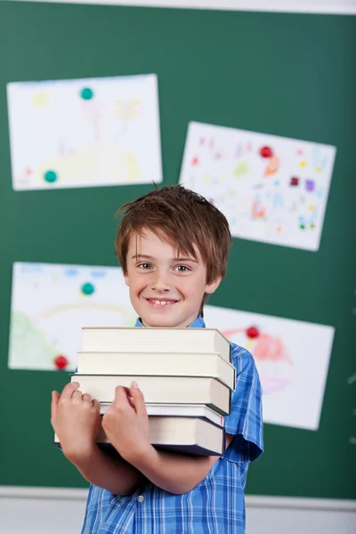Jovem estudante elementar segurando uma pilha de livros — Fotografia de Stock