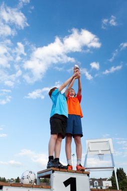 Boys Holding Trophy While Standing On Winners Podium Against Sky clipart