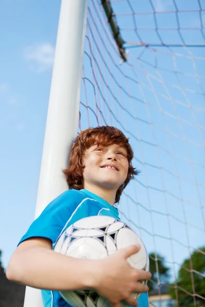 Kleiner Junge hält Fußball, während er sich an Netzstange lehnt — Stockfoto