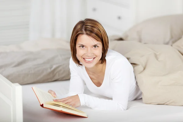 Mulher dando um sorriso enquanto lê um livro na cama — Fotografia de Stock