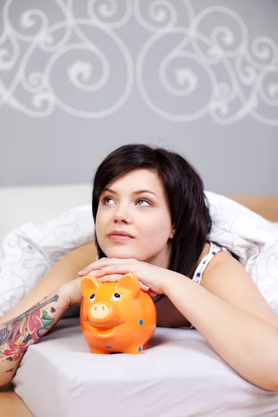 Mujer joven con Piggybank mirando hacia arriba mientras se acuesta en la cama — Foto de Stock
