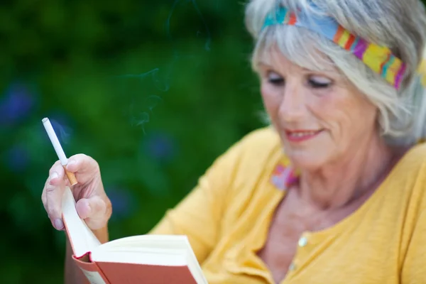 Seniorin liest Buch während sie Zigarette raucht — Stockfoto