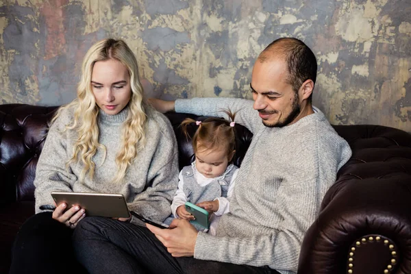 Busy parents hold gadgets, sit on couch with cute baby girl watch cartoon together at home. Soft focus