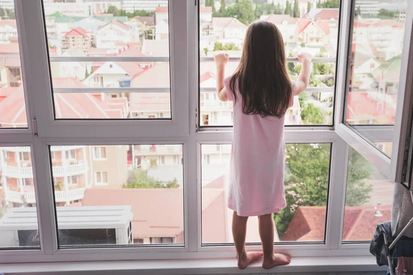 Uma criança está perigosamente parada em uma janela aberta. Crianças e janelas abertas. Uma rapariga de pijama nocturno está ao pé da janela. Segurança das crianças em casa — Fotografia de Stock