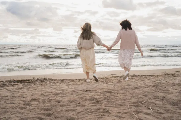Két lány kéz a kézben megy a tengerhez. Sétálj az óceán mellett ősszel meleg ruhában. A nők közötti kapcsolatok. — Stock Fotó