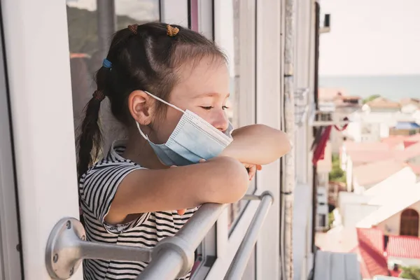 Egy kislány megbetegedett a nyaraláson. A lánynak hiányzik az otthona a betegség miatt. Gyerekek orvosi maszkban. Egy lány néz ki az ablakon, ahogy a többi gyerek sétál. — Stock Fotó
