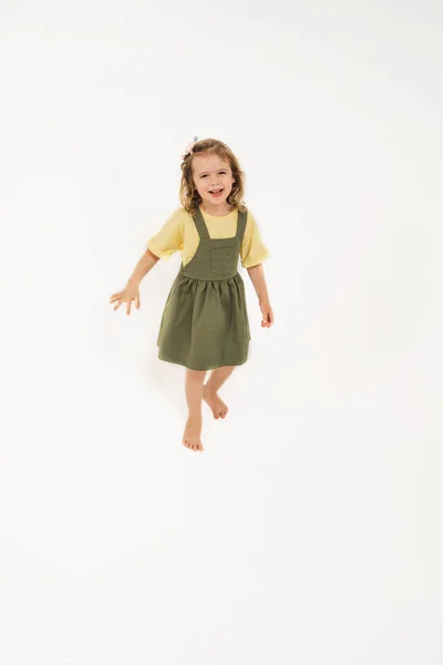 Ein Mädchen in einer grünen Uniform. Ein schönes Mädchen jubelt und springt. Ein Kind mit blonden Haaren auf hellem Hintergrund. Echte Kindergefühle bei einem Kind. — Stockfoto