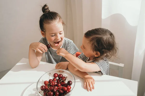 Starsza siostra karmi młodszą jagodami. Pyszne letnie jagody. Dzieci chętnie jedzą soczyste jagody. Dwie siostry w koszulkach w paski jedzą — Zdjęcie stockowe