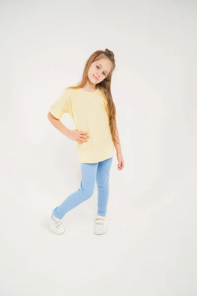 Ребенок в жёлтой футболке и синих штанах. Ребёнок с длинными светлыми волосами. Девушка с модельной внешностью на светлом фоне — стоковое фото
