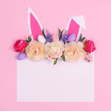 Yaratıcı Paskalya tebrik kartı. Kağıt kart fotokopi alanı tavşan kulakları ve pastel pembe arka planda bahar çiçekleriyle dekore edilmiş. Düz çiçek konsepti.