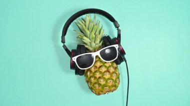 Güneş gözlüklü ve kulaklıklı yaratıcı ananas dans ediyor ve cyan arka planda zıplıyor. Dur hareketi düz yatıyordu