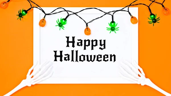 Happy Halloween text in white frame with skeleton hands and laterns on orange background. Flat lay
