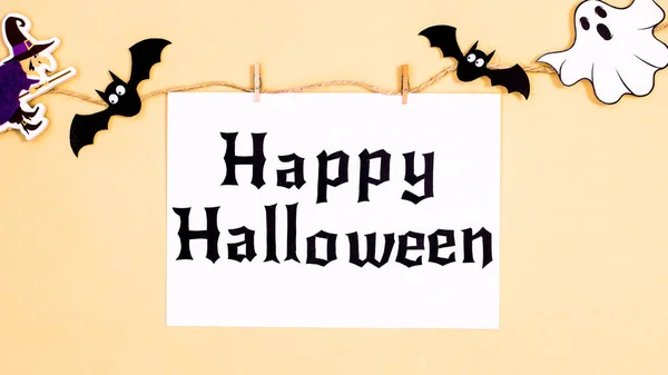 Creative paper card with Happy Halloween text hanged on rope with scary stickers for Halloween on beige  background. Flat lay