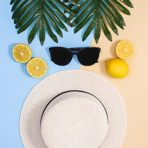 Summer beach concept with hat, lemons, sunglasses and natural palm leaves on blue and beige background. Flat lay