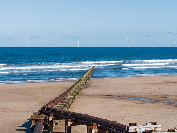 Outfall pipe at Cambois, Northumberland, UK into blue sea with wind turbines in the distance.