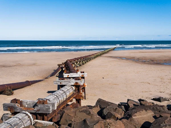 Outfall pipe at Cambois, Northumberland, UK into blue sea with wind turbines in the distance.